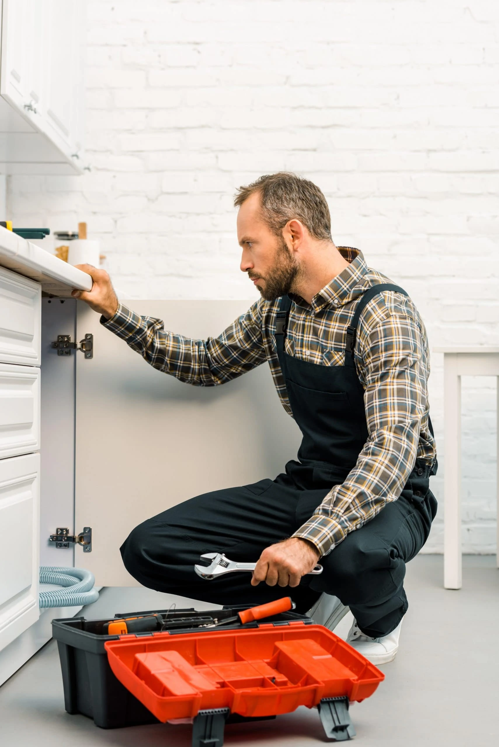 side view of handsome repairman holding adjustable wrench and looking under broken sink in kitchen